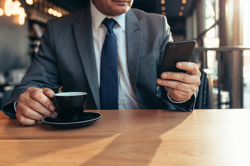 Abgeschnittene Aufnahme eines Smartphones in der Hand eines älteren Geschäftsmannes mit Kaffee auf einem Cafétisch. Älterer Geschäftsmann schreibt eine SMS auf seinem Handy in einem Café. - JLPSF22049