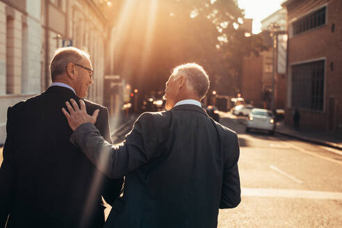 Rückansicht von zwei Geschäftsleuten, die nach einem erfolgreichen Geschäftstreffen im Freien spazieren gehen und sich neben einem Bürogebäude unterhalten. Ältere Geschäftsleute gehen an einem sonnigen Tag zusammen spazieren. - JLPSF22052