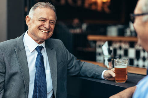 Lächelnder älterer Geschäftsmann mit einem Glas Bier im Gespräch mit einem Kollegen an der Bar nach der Arbeit. Geschäftsleute entspannen sich nach der Arbeit an der Bar. - JLPSF22055