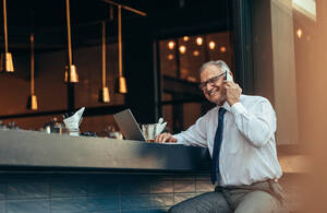 Lächelnder älterer Mann in formeller Kleidung, der an einem Bartresen mit Laptop sitzt und mit seinem Handy telefoniert. - JLPSF22059