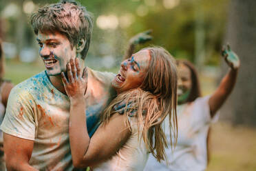 Lächelnde Frau, die ihrem Freund Holi-Farbe auf das Gesicht aufträgt. Freunde genießen und haben Spaß beim Holi-Spielen in einem Park. - JLPSF22074
