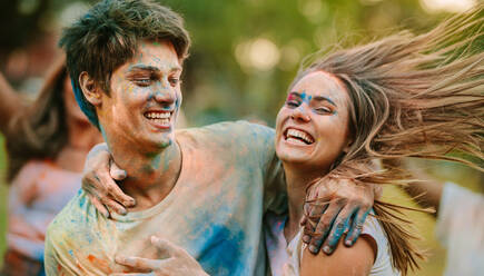 Man and woman with their faces smeared with holi colours having fun outdoors. Happy couple holding each other playing holi with their friends in a park. - JLPSF22075
