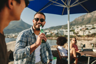 Lächelnder Mann mit Sonnenbrille, der Bier trinkt und sich mit seinem Freund unterhält, mit zwei Frauen im Hintergrund. Männer im Urlaub in der Nähe eines Strandes trinken Wein. - JLPSF22080