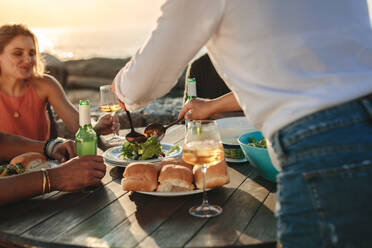 Freunde sitzen an einem Tisch in der Nähe des Meeres und essen Snacks und Getränke. Frau genießt Essen und Getränke im Freien mit Freunden an einem sonnigen Abend. - JLPSF22084