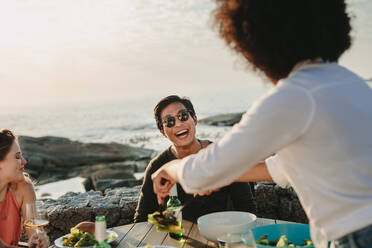 Multiethnische Freunde sitzen an einem Esstisch in der Nähe des Strandes mit Getränken und Snacks. Mann mit Sonnenbrille sitzt lachend am Strandtisch mit Freunden und genießt Essen und Getränke. - JLPSF22085