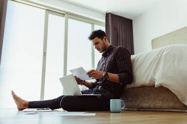 Man working on laptop computer sitting on the floor. Freelancer looking at a paper while working on laptop computer at home. - JLPSF22086