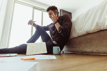 Man working on laptop computer sitting on the floor at home. Freelancer sitting on floor beside a bed and talking over mobile phone looking at his diary with a coffee cup by his side. - JLPSF22092