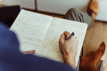 Rear view close up of man sitting on floor at home and making notes in diary. Close up of hand of man holding a pen and writing in diary. - JLPSF22101