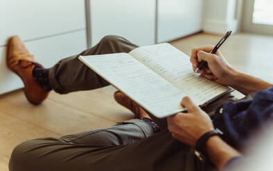 Seitenansicht eines Mannes, der zu Hause auf dem Boden sitzt und sich Notizen in sein Tagebuch macht. Nahaufnahme eines Mannes, der einen Stift hält und in sein Tagebuch schreibt. - JLPSF22102