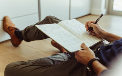 Side view of man sitting of floor at home and making notes in diary. Close up of man holding a pen and writing in diary. - JLPSF22102