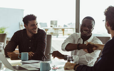 Businessmen discussing work sitting at conference table in office. Men shaking hands and smiling during a business meeting. - JLPSF22108