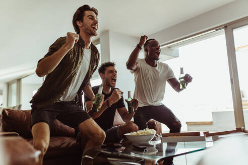 Drei Freunde springen aufgeregt mit Bierflaschen in der Hand herum, während sie ein Sportspiel im Fernsehen verfolgen. - JLPSF22115