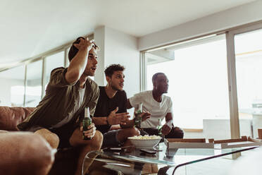 Drei Freunde sehen sich ein Spiel im Fernsehen an und halten Bierflaschen in der Hand. Männer sitzen mit Getränken und Snacks auf dem Sofa und sehen zu Hause fern. - JLPSF22116