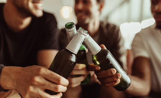 Nahaufnahme von drei Freunden, die bei einer Party zu Hause mit ihren Bierflaschen anstoßen. Männer, die zu Hause sitzend mit ihren Flaschen anstoßen. - JLPSF22121