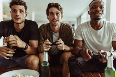 Friends having fun with drinks and snacks while playing video game at home. Two men playing video game holding joysticks while another man watches with a bottle of beer in hand. - JLPSF22122