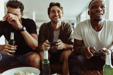 Friends having fun with drinks and snacks while playing video game at home. Two men playing video game holding joysticks while another man watches with a bottle of beer in hand. - JLPSF22123