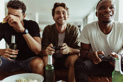 Zwei Männer spielen ein Videospiel und halten Joysticks, während ein anderer Mann mit einer Bierflasche in der Hand zusieht. - JLPSF22123