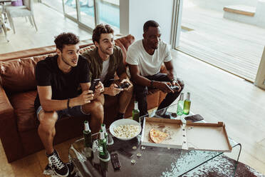 Freunde haben Spaß mit Getränken und Snacks, während sie zu Hause ein Videospiel spielen. Zwei Männer spielen ein Videospiel und halten Joysticks, während ein anderer Mann ein Handy in der Hand hält. - JLPSF22125
