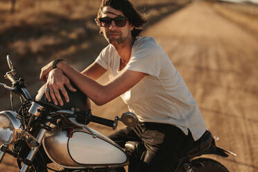 Porträt eines gutaussehenden jungen Mannes auf seinem Motorrad auf einer Landstraße. Männlicher Biker auf seinem Motorrad sitzend und entspannt auf der Landstraße. - JLPSF22137