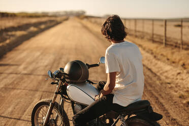 Rückansicht eines jungen Mannes, der auf seinem Motorrad sitzt und auf eine leere Landstraße schaut. Junger Motorradfahrer, der sich auf seinem Motorrad entspannt und auf die Landstraße schaut. - JLPSF22138