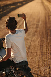 Rückansicht eines jungen Mannes, der ein Selfie mit seinem Smartphone macht, während er auf seinem Motorrad auf einer Landstraße sitzt. Ein Motorradfahrer, der Erinnerungen an seine Fahrt auf dem Land festhält. - JLPSF22139