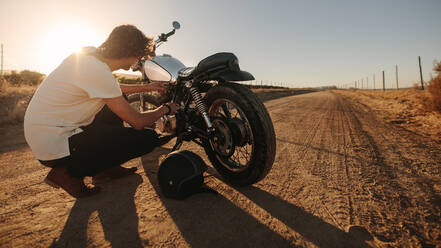 Junger Mann überprüft die Kraftstoffleitung seines Motorrads auf einer Landstraße. Motorradfahrer repariert sein Motorrad auf einer leeren Landstraße. - JLPSF22140