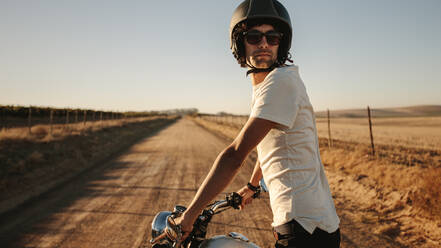 Young man wearing a helmet and sunglasses standing with his bike and looking back on rural road. Male bike on countryside road. - JLPSF22141