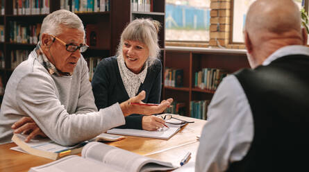 Zwei ältere Männer und eine Frau sitzen in einer Bibliothek mit Kursbüchern auf dem Tisch und diskutieren. Ältere Männer und eine Frau tauschen Ideen aus und diskutieren in einer Bibliothek sitzend. - JLPSF22153