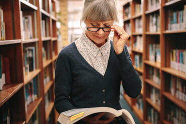 Elderly woman standing in a library holding a book. Senior woman looking at a book in a library. - JLPSF22156