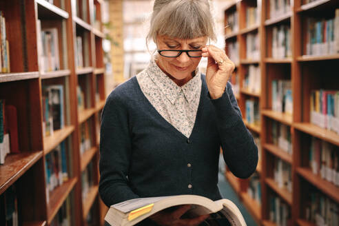 Ältere Frau steht in einer Bibliothek und hält ein Buch in der Hand. Ältere Frau betrachtet ein Buch in einer Bibliothek. - JLPSF22156