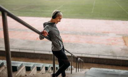 Smiling woman in fitness clothes standing on the stairs of a stadium listening to music. Woman standing on the stairs taking support of the railing in the stands of a track and field stadium. - JLPSF22157