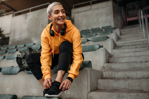 Fitness-Frau sitzt auf der Tribüne eines Stadions und bindet Schnürsenkel. Fröhliche Sportlerin, die sich auf ihr Training vorbereitet und ihre Schuhe trägt. - JLPSF22167