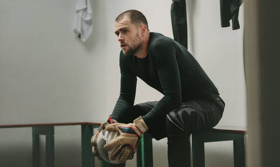 Goalkeeper sitting in a room holding a football and thinking. Soccer player sitting on bench inside the dressing room looking away. - JLPSF22169