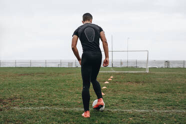 Rückansicht eines Fußballspielers, der mit Hilfe von auf dem Feld aufgestellten Kegeln das Dribbeln übt. Fußballspieler, der früh am Morgen auf dem Feld übt. - JLPSF22173