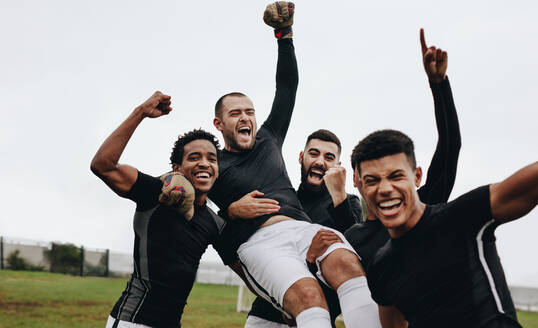 Group of happy soccer players celebrating a win by lifting their goalkeeper. Footballers celebrating victory by raising their hand in air and screaming in excitement. - JLPSF22188