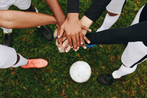 Top view of hands of players placed one over the other standing in a huddle. Players standing in a huddle joining their hands together in the centre with a soccer ball on the ground. - JLPSF22197