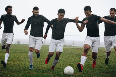 Fußball spielende Männer, die an einem regnerischen Morgen einem Fußball hinterherlaufen. Fußballer, die versuchen, den Ball zu erobern, laufen auf dem Fußballfeld. - JLPSF22199