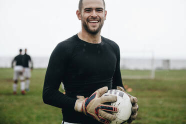 Porträt eines fröhlichen Torwarts, der einen Fußball in der Hand hält. Fußballspieler, der auf dem Feld steht und einen Fußball hält und am Morgen übt. - JLPSF22202