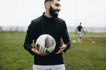 Lächelnder Fußballspieler, der während des Trainings auf dem Feld steht und einen Fußball hält. Fröhlicher Fußballspieler, der am Morgen auf dem Feld übt, während seine Mannschaftskameraden im Hintergrund spielen. - JLPSF22205