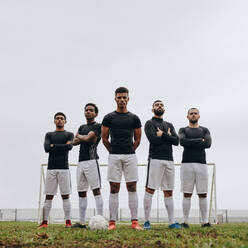 Tiefblick auf Fußballer, die während des Trainings an einem bewölkten Tag auf einem Fußballplatz stehen. Fünf Fußballer stehen mit verschränkten Armen auf einem Fußballplatz zusammen. - JLPSF22212