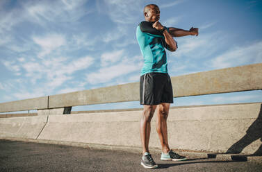 Afroamerikanischer Mann in Sportkleidung, der seine Arme ausstreckt. Männlicher Sportler beim Aufwärmen vor einem Lauf am Morgen in der Stadt. - JLPSF22217