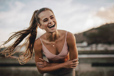 Lächelnde Läuferin, die am Morgen im Freien steht. Frau in Sportkleidung lacht mit verschränkten Armen und ruht sich nach dem Training aus. - JLPSF22224