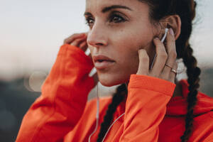 Close up of female athlete with earphones standing outdoors. Fitness woman taking break after morning run and listening to music with earphones. - JLPSF22232