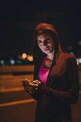 Fitte junge Frau in Kapuzenshirt steht mit Handy im Freien. Läuferin macht Pause nach dem Training im Freien bei Nacht. - JLPSF22242