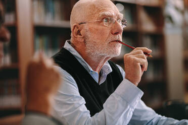 Elderly man sitting in classroom listening to a lecture with concentration. Senior man sitting in a library with pencil to his mouth and thinking. - JLPSF22250