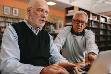 Zwei ältere Männer sitzen in einem Klassenzimmer und arbeiten gemeinsam an einem Laptop. Ein älterer Mann sitzt in einer Bibliothek und arbeitet mit einem Bleistift im Mund an einem Laptop mit seinem Klassenkameraden. - JLPSF22260