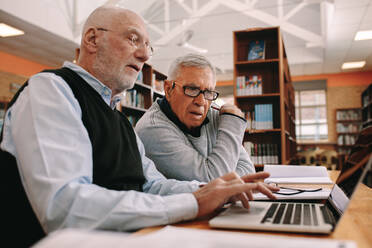 Seitenansicht von zwei älteren Männern, die in einem Klassenzimmer an einem Laptop arbeiten. Ältere Männer sitzen in einer Bibliothek und lernen ihre Kurse an einem Laptop. - JLPSF22261