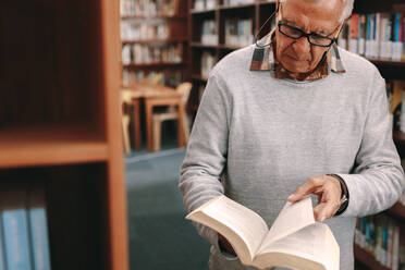 Nahaufnahme eines älteren Herrn, der in einer Bibliothek steht und in einem Lehrbuch blättert. Älterer Mann, der in einer Universitätsbibliothek nach Nachschlagewerken sucht. - JLPSF22265