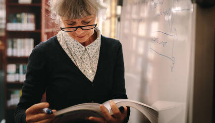 Nahaufnahme einer älteren Frau, die in einer Bibliothek steht und in einem Lehrbuch blättert. Ältere Frau, die in einer Universitätsbibliothek nach Nachschlagewerken sucht. - JLPSF22269
