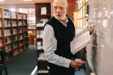 Ein älterer Dozent, der in einer Klasse unterrichtet, schreibt auf einer Tafel und hält ein Buch in der Hand. Ein Professor, der sich umdreht, während er an der Tafel schreibt, steht im Klassenzimmer. - JLPSF22271